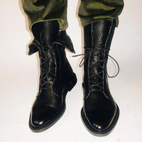 Bottes en cuir noir à lacets croisés, bout pointu, portées avec pantalon vert.