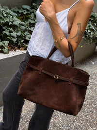 Sac en daim marron porté en bandoulière, anses courtes et boucle décorative.