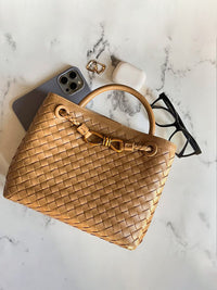 Sac tressé beige avec anse rigide, fermoir décoratif doré et téléphone visible.