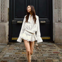 Robe courte blanche plissée, col V, manches longues, ceinture nouée, jupe volantée
